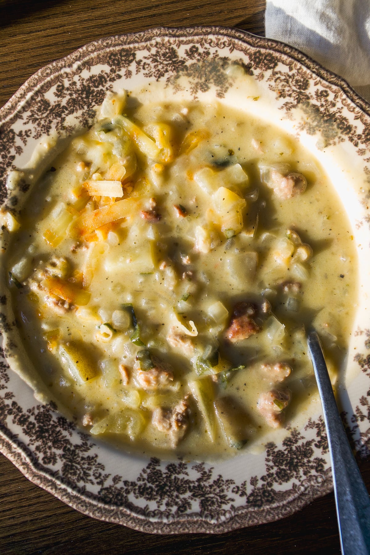 A bowl of creamy vegetable and sausage soup served in a patterned porcelain bowl with a spoon.