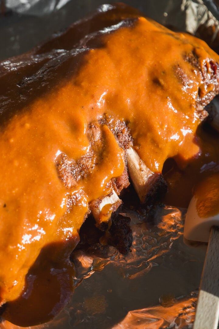 A rack of ribs coated in thick, orange barbecue sauce on a foil-lined tray.