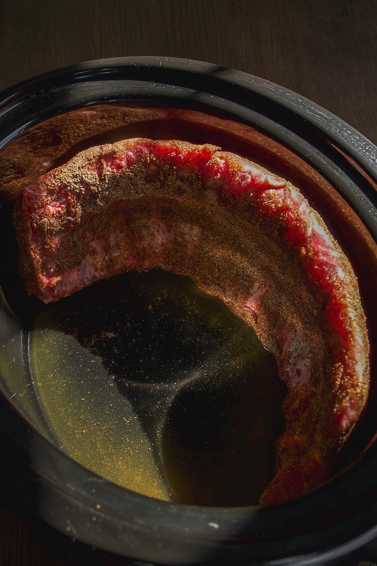 A rack of raw ribs coated in dry spice rub sitting in a slow cooker with liquid at the bottom.