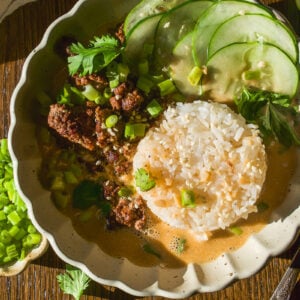 A bowl of ground meat and rice in a creamy sauce, topped with sliced cucumbers, green onions, and cilantro.