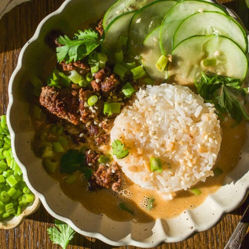 A bowl of ground meat and rice in a creamy sauce, topped with sliced cucumbers, green onions, and cilantro.