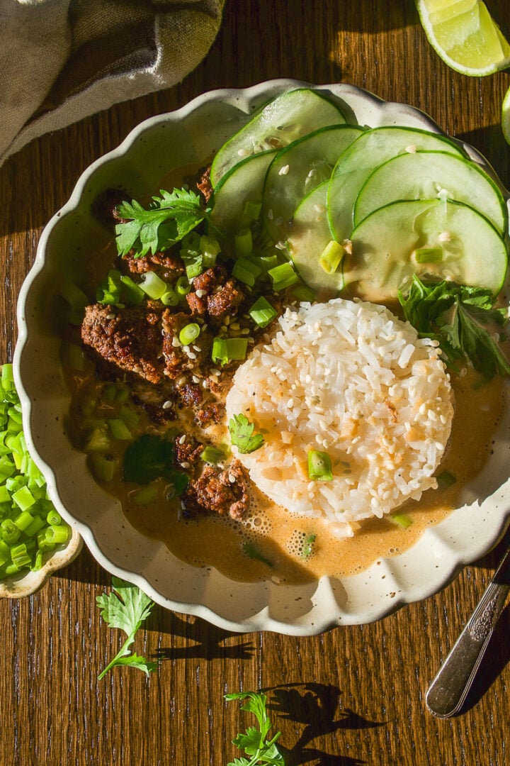 A bowl of ground meat and rice in a creamy sauce, topped with sliced cucumbers, green onions, and cilantro.