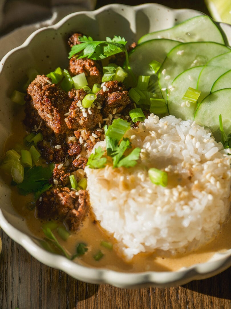 A bowl containing ground meat, a mound of white rice, and cucumber slices, garnished with scallions, cilantro, and sesame seeds in a creamy sauce.