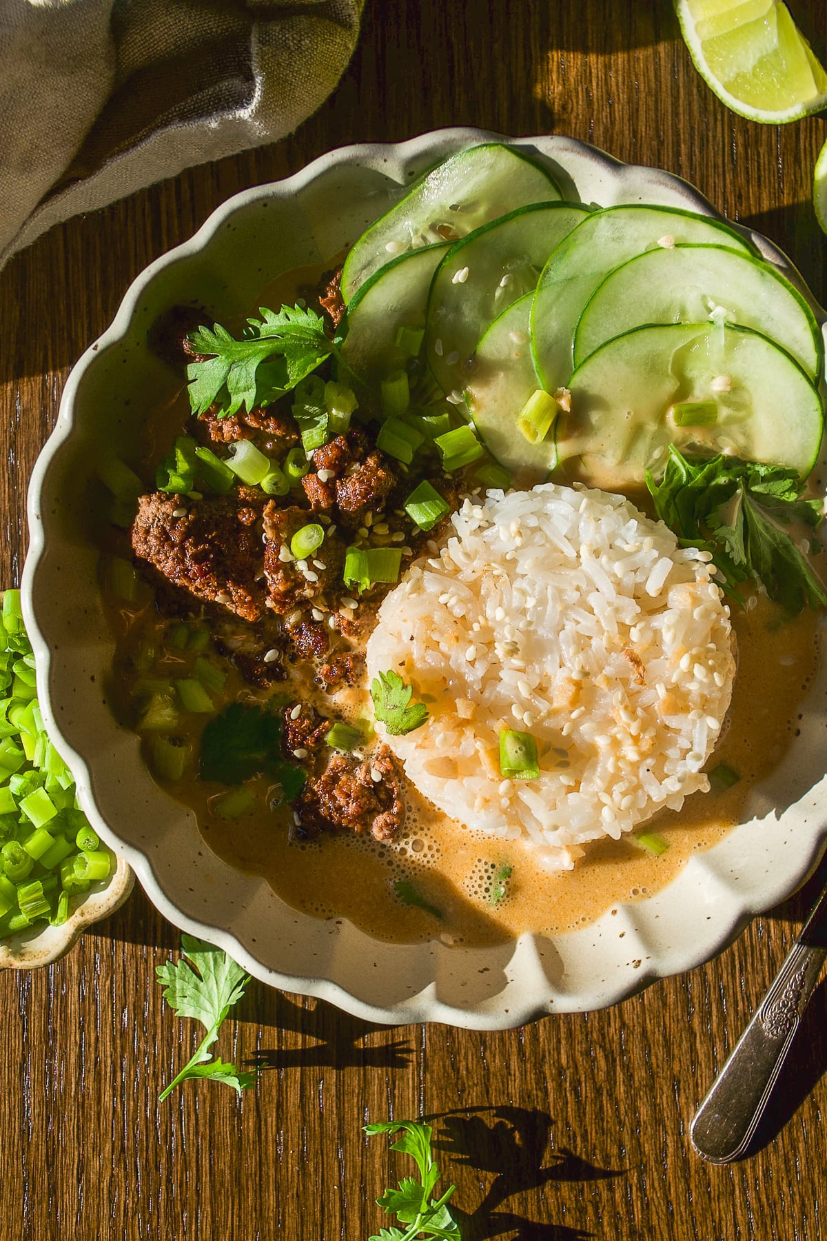 A bowl of curry topped with ground meat, a scoop of white rice, sliced cucumbers, and cilantro.