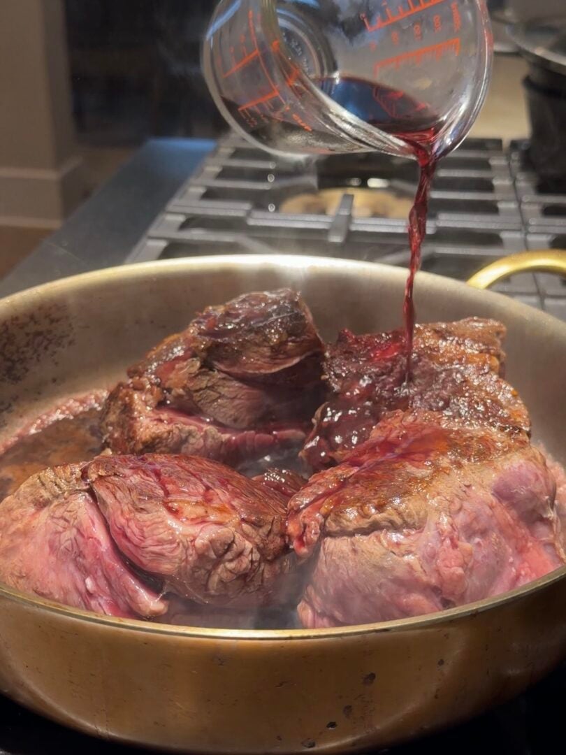 Red wine being poured from a measuring cup onto seared beef steaks in a metal pan.