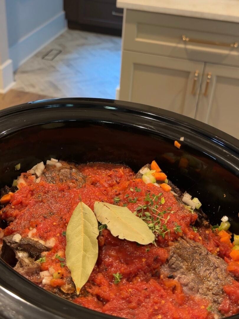Beef, carrots, onions, and herbs simmering in a tomato-based sauce in a slow cooker, topped with two bay leaves.
