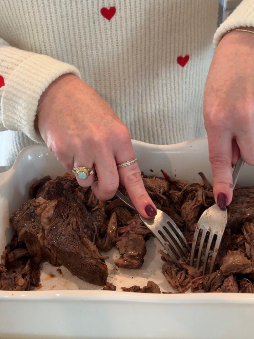 Hands shredding cooked roast beef in a white baking dish with two forks.