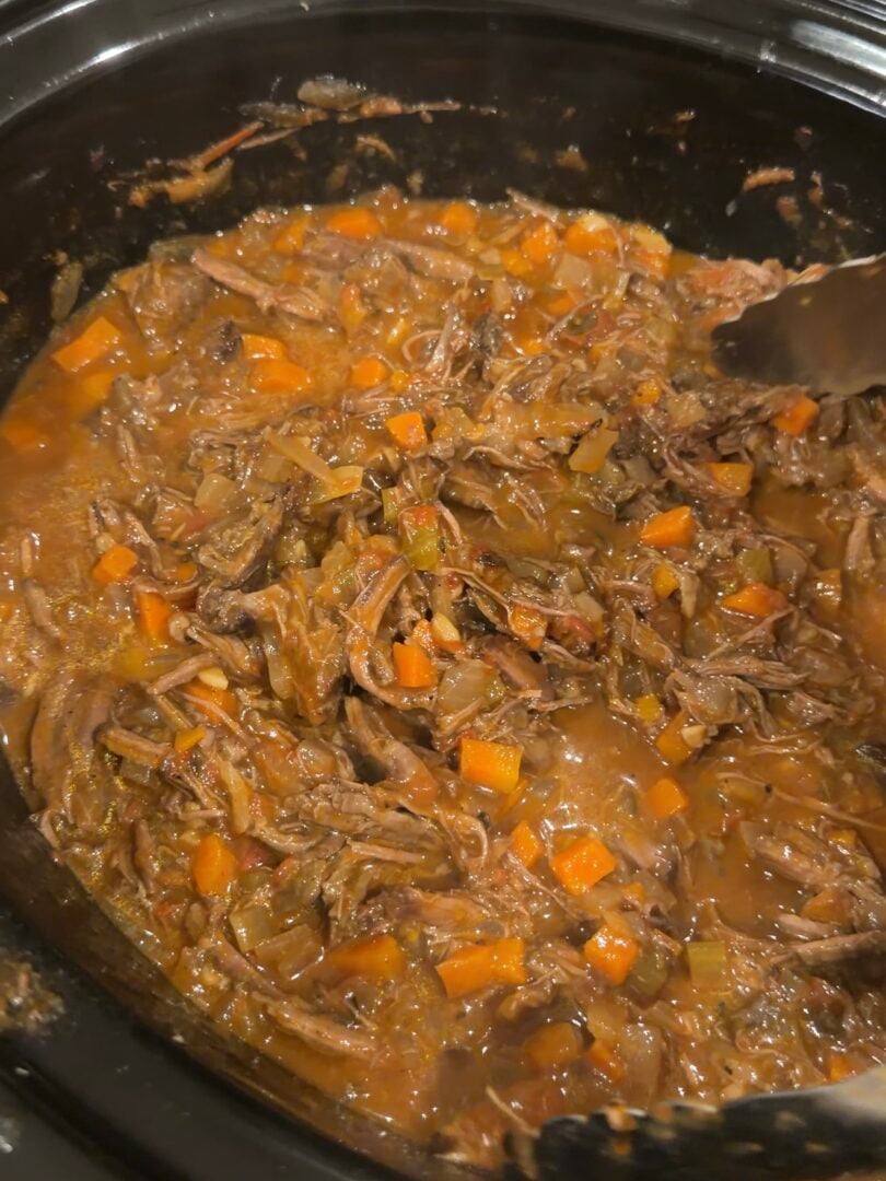 Shredded beef stew with diced carrots and onions in a slow cooker, being stirred with metal tongs.