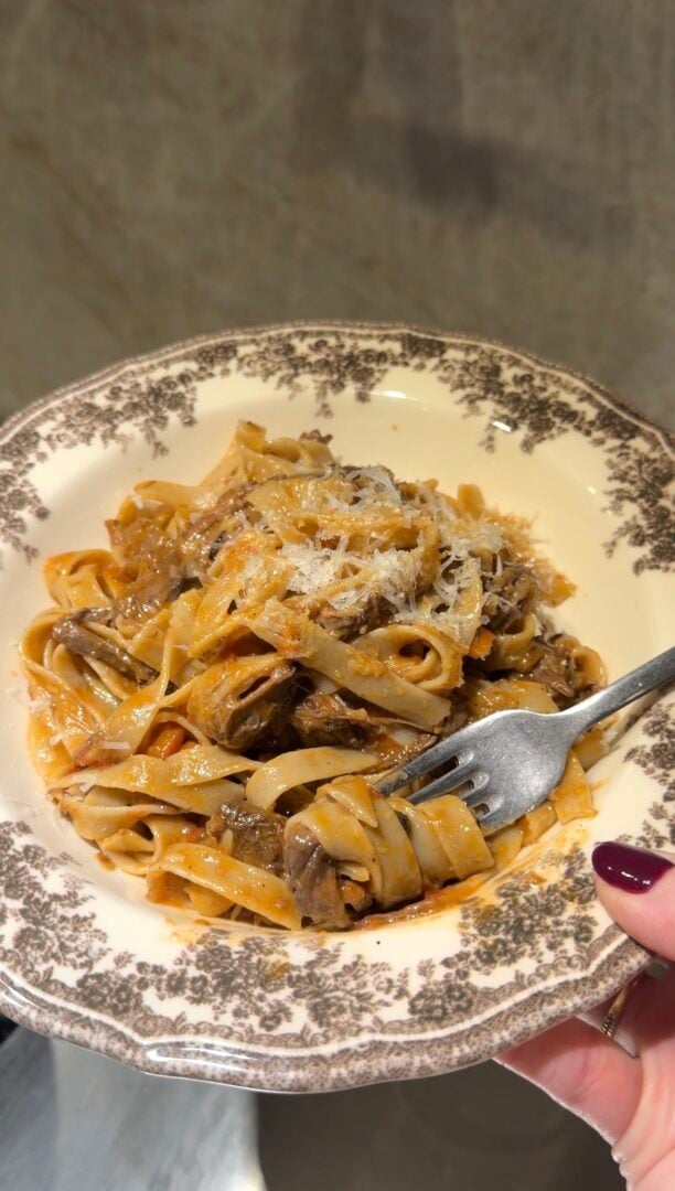 A fork twirls a portion of beef ragu tagliatelle topped with grated cheese in a decorative bowl.