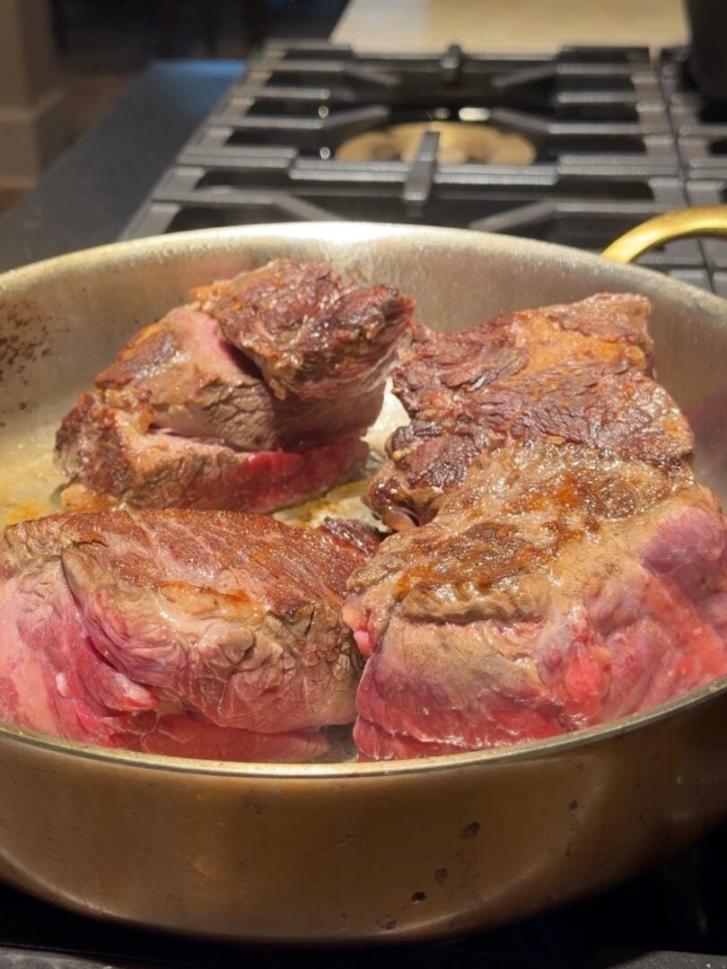 Four thick pieces of seared steak browning in a metal skillet on a stovetop.