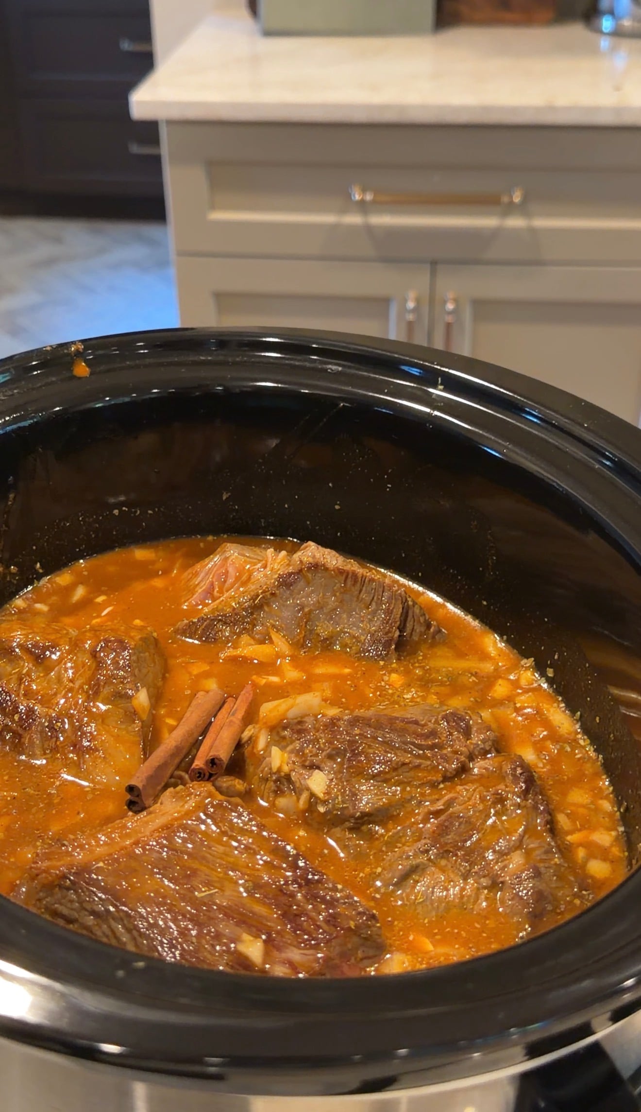 Chunks of beef simmer in a slow cooker filled with red sauce, onions, and visible cinnamon sticks. The slow cooker sits on a kitchen counter with cabinets in the background.