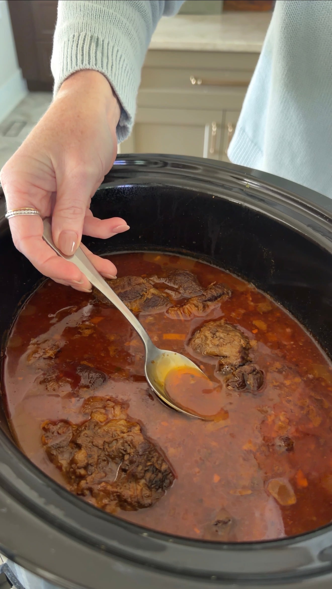 A hand holding a spoon hovers over a slow cooker filled with a rich, red stew and chunks of braised meat.