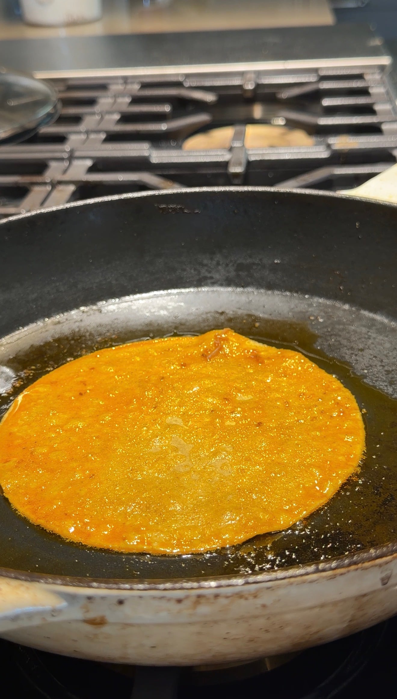 A round, orange tortilla is frying in oil on a black pan over a stove, with bubbling edges and a golden hue.