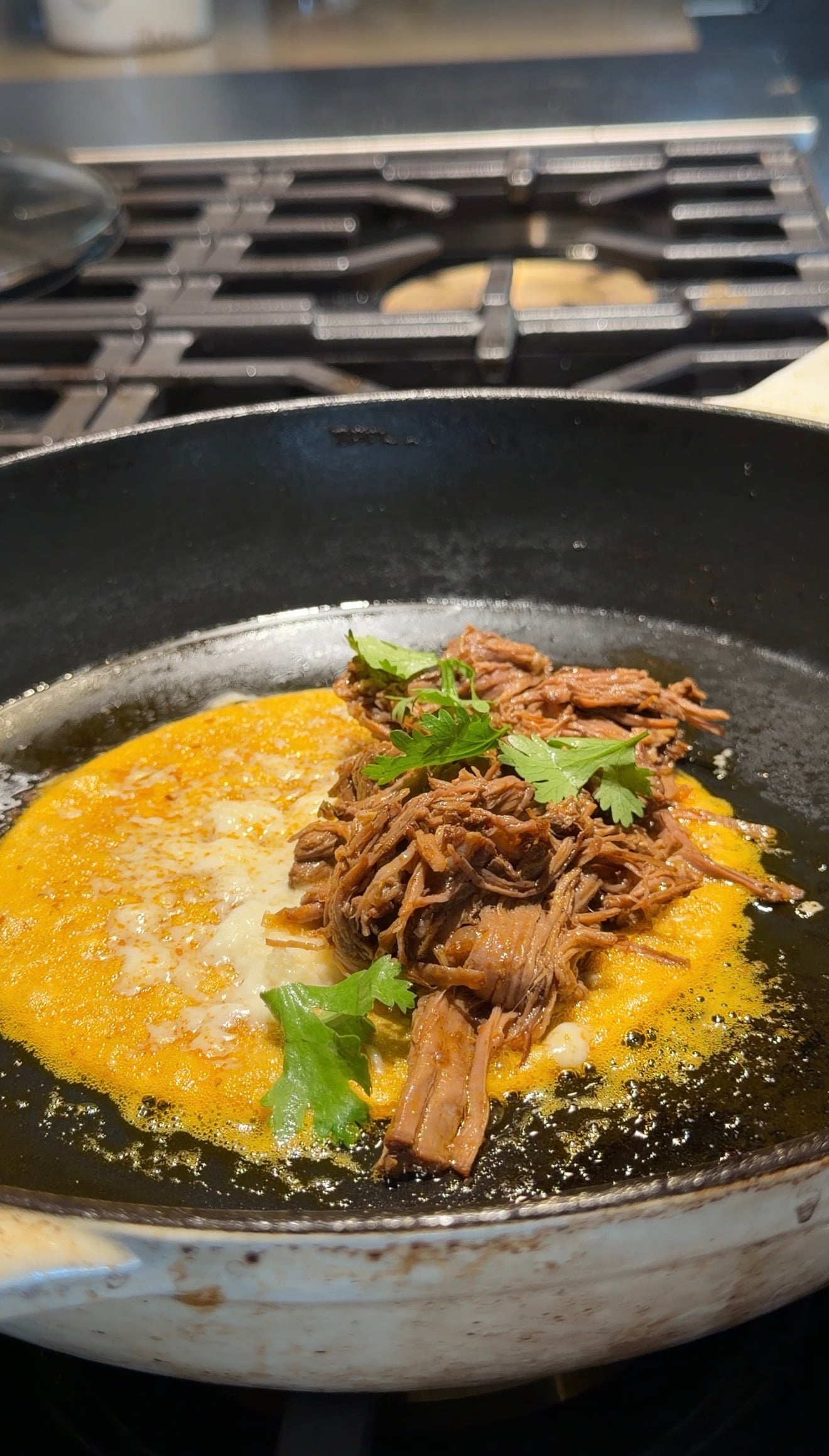 Shredded beef and fresh cilantro topping a yellow omelet cooking in a black skillet on a stovetop.