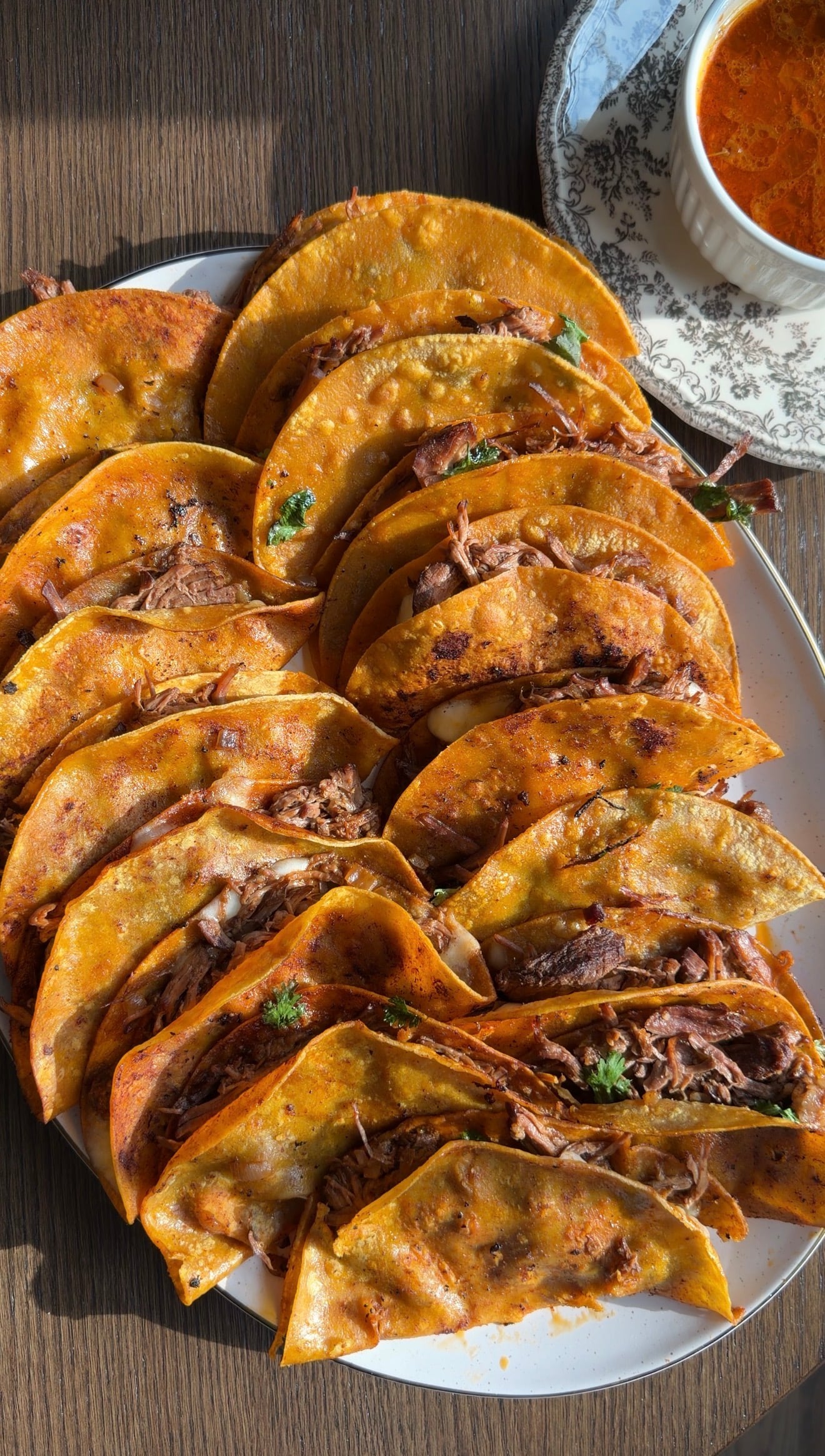 A plate filled with golden, crispy birria tacos stuffed with shredded meat, garnished with herbs, sits next to a small bowl of dipping broth on a wooden table.