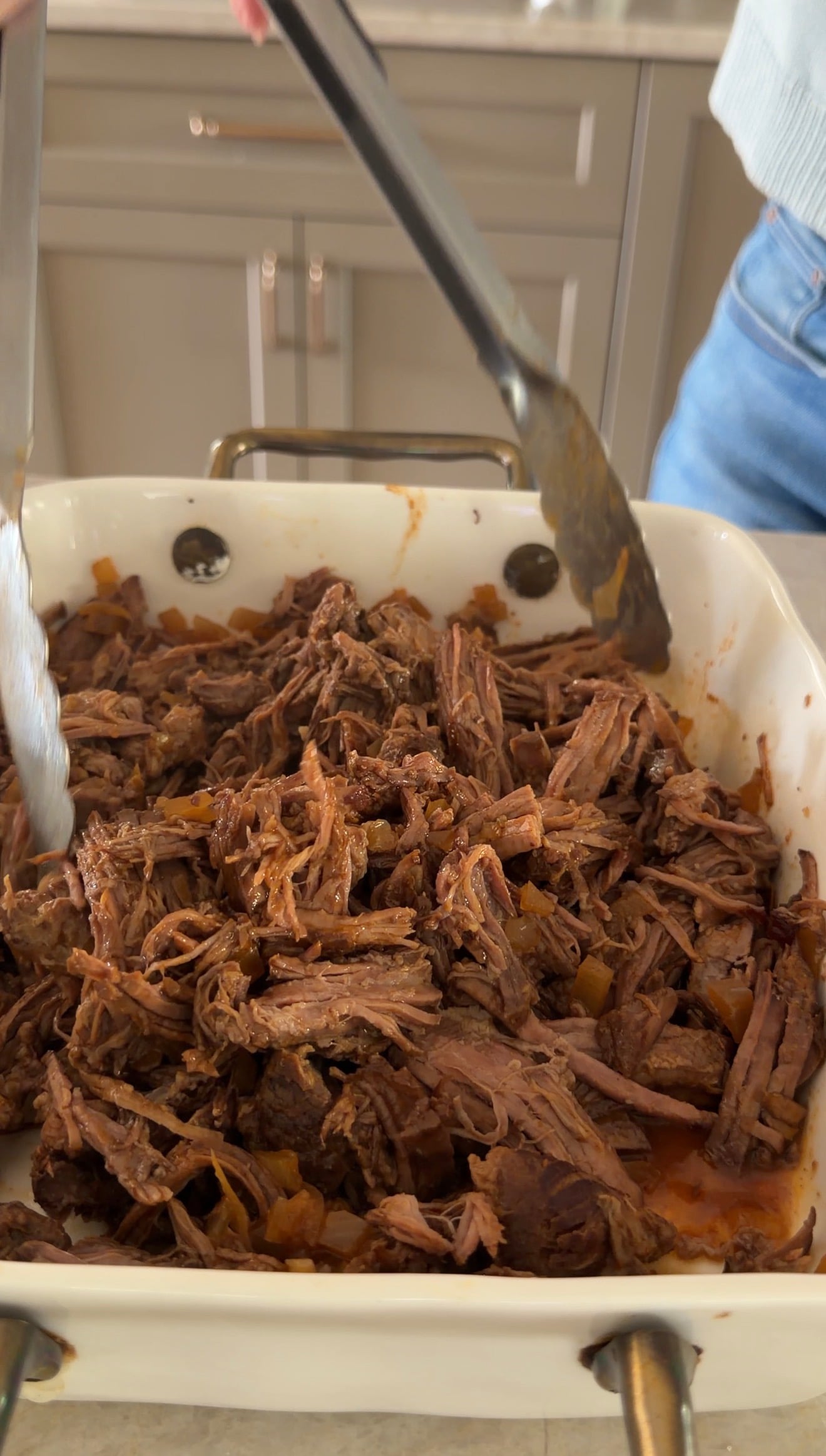 A person uses metal tongs to serve shredded beef from a white rectangular dish. The beef appears tender and cooked in a savory sauce. The background shows part of a person in blue jeans and a kitchen counter.