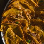 A close-up of several golden, crispy tacos filled with shredded meat, garnished with herbs, arranged on a white plate in sunlight.