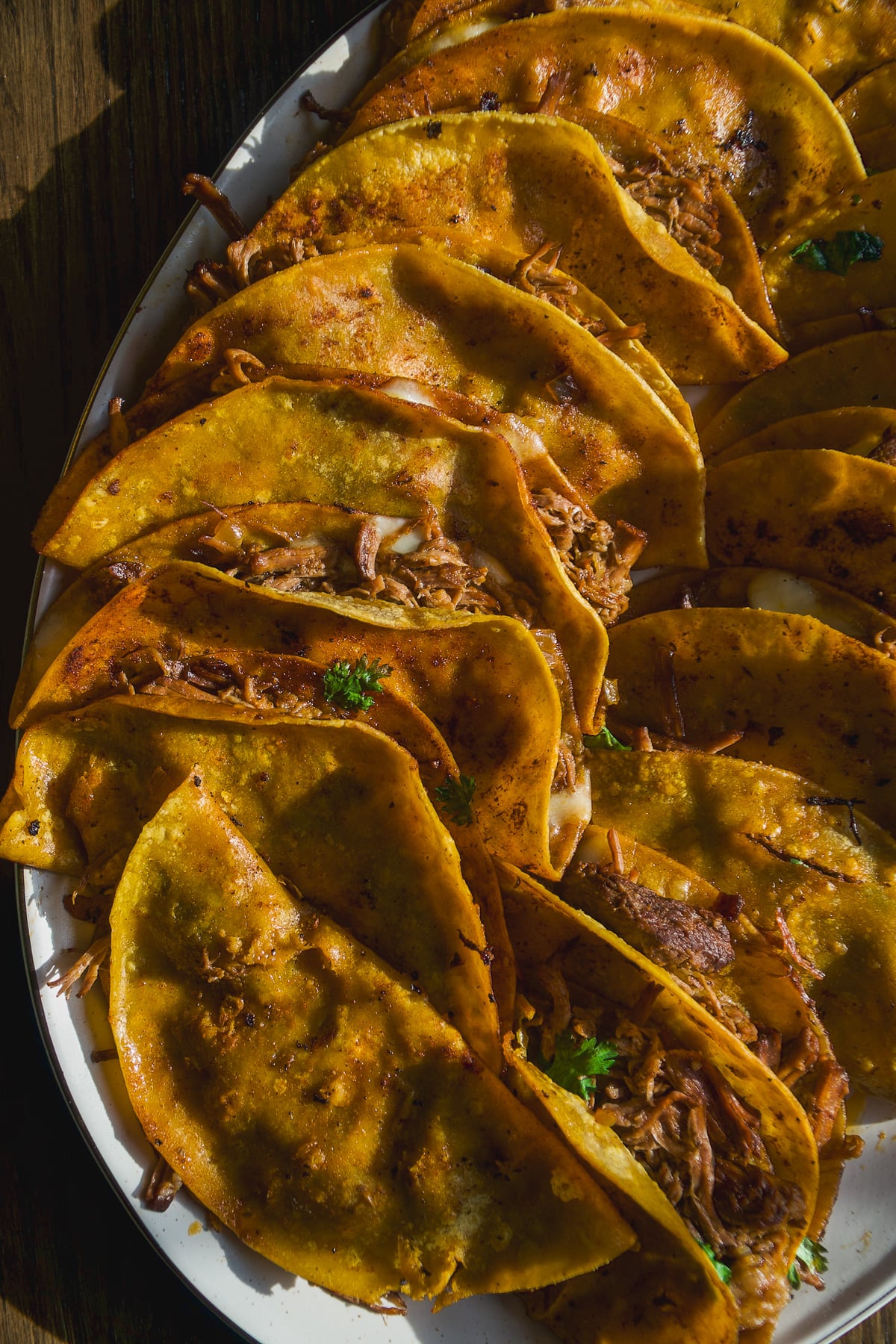 A close-up of several golden, crispy tacos filled with shredded meat, garnished with herbs, arranged on a white plate in sunlight.
