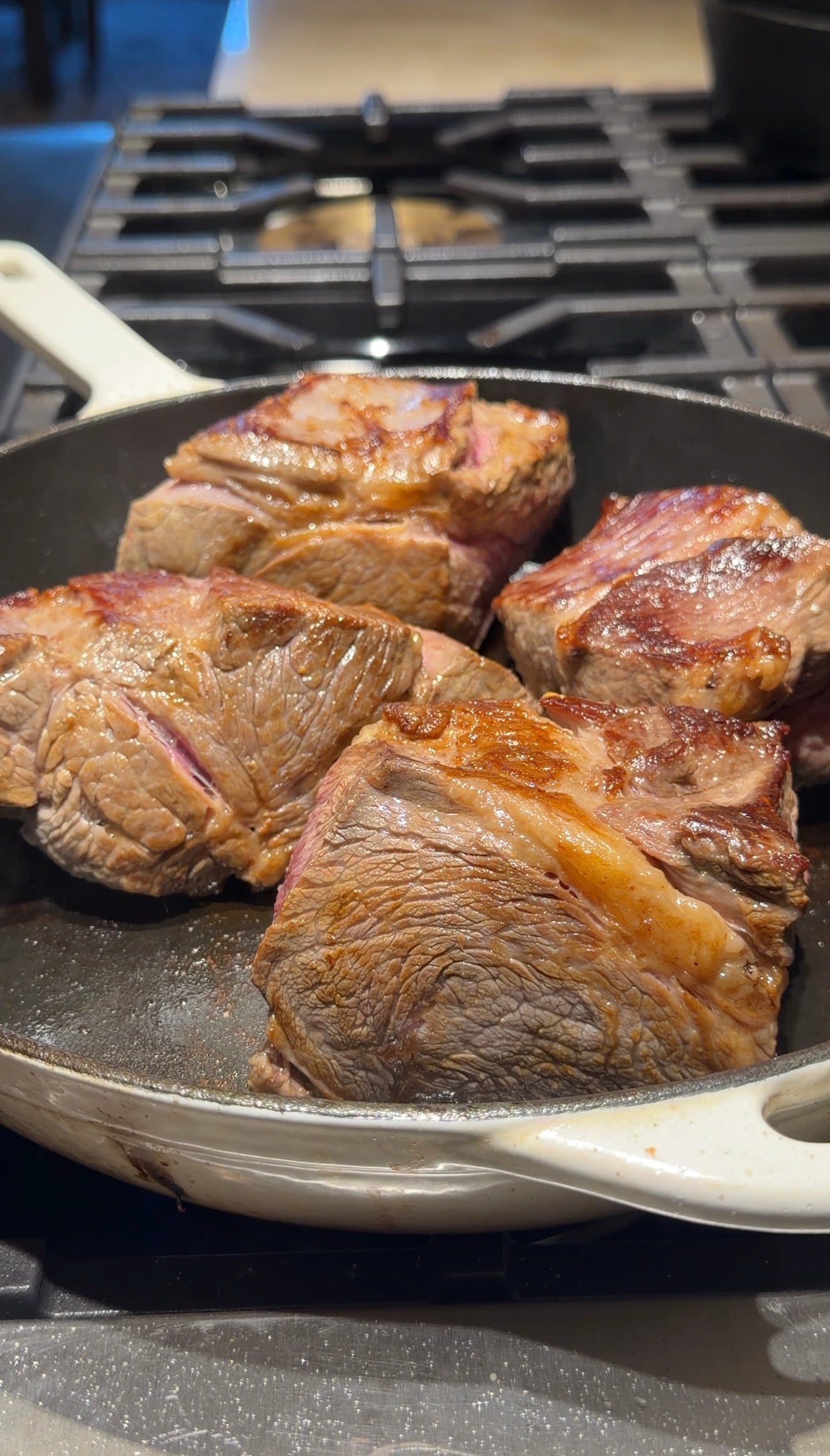 Four large pieces of seared beef are cooking in a black skillet on a stovetop. The meat has a browned, caramelized surface and is arranged closely together in the pan.