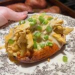 A baked sweet potato topped with creamy shredded chicken and chopped green onions on a patterned plate.