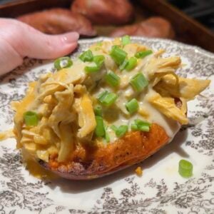 A baked sweet potato topped with creamy shredded chicken and chopped green onions on a patterned plate.