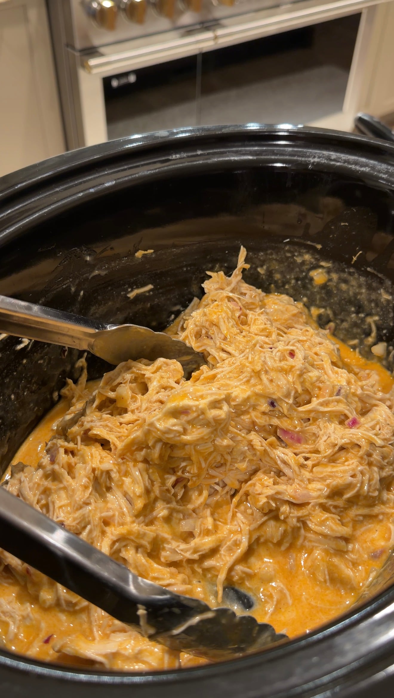 Shredded chicken in a creamy orange sauce inside a slow cooker with metal tongs.