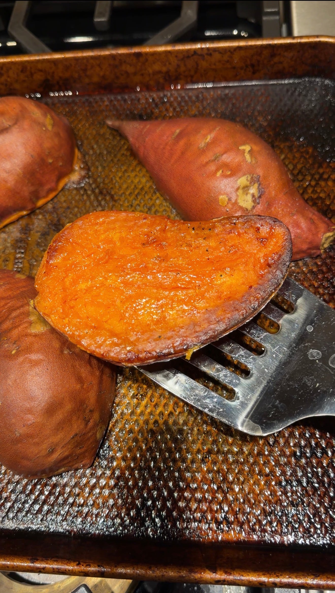 A metal spatula lifting a slice of roasted sweet potato from a baking sheet with other roasted sweet potatoes.