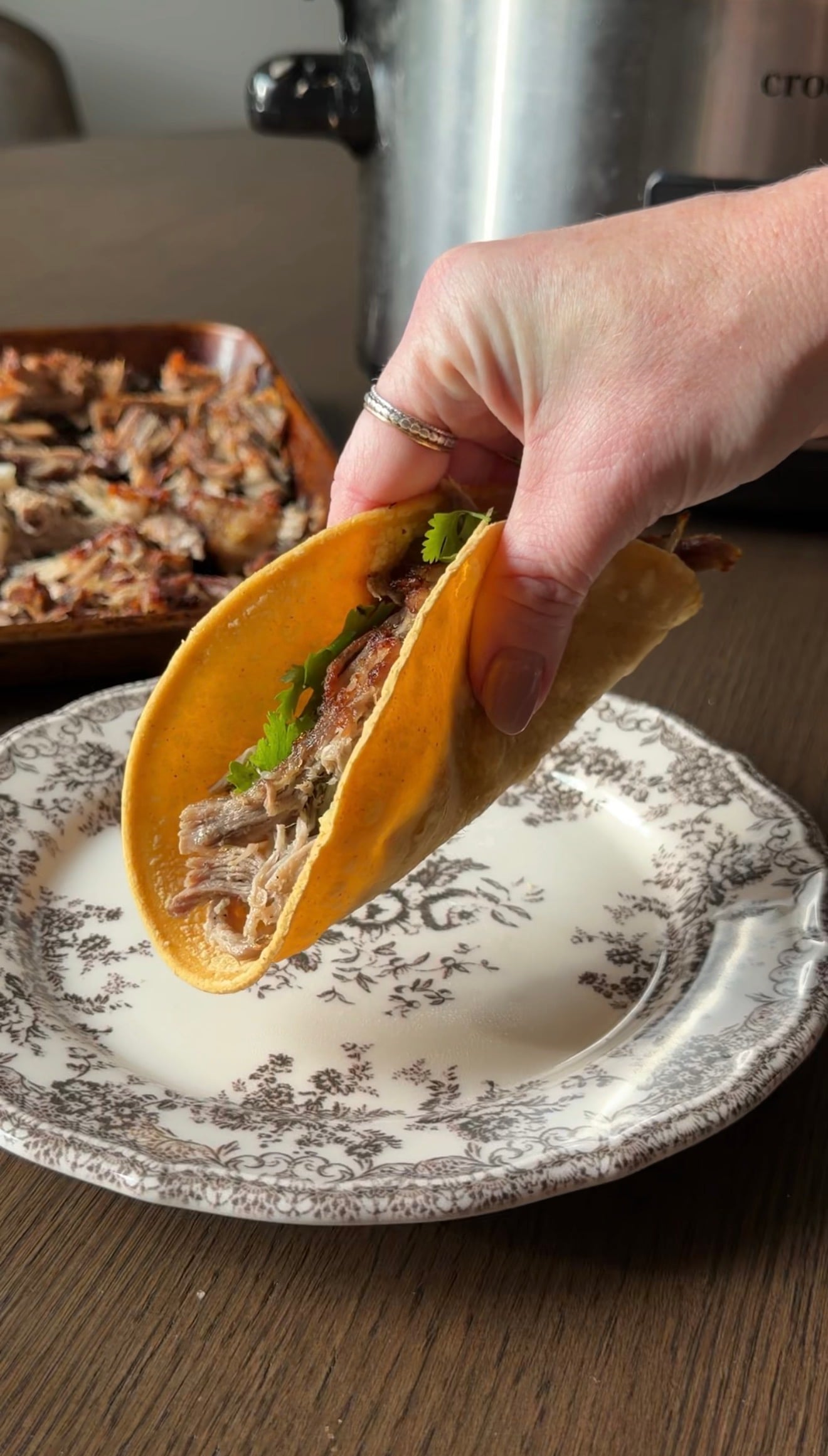 A hand holds a crunchy taco filled with shredded meat and cilantro over a decorative plate.