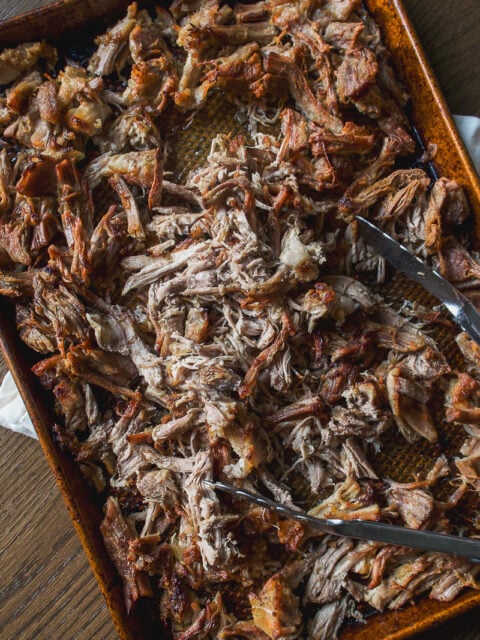 A metal baking sheet filled with shredded crockpot carnitas, with metal tongs resting on top.