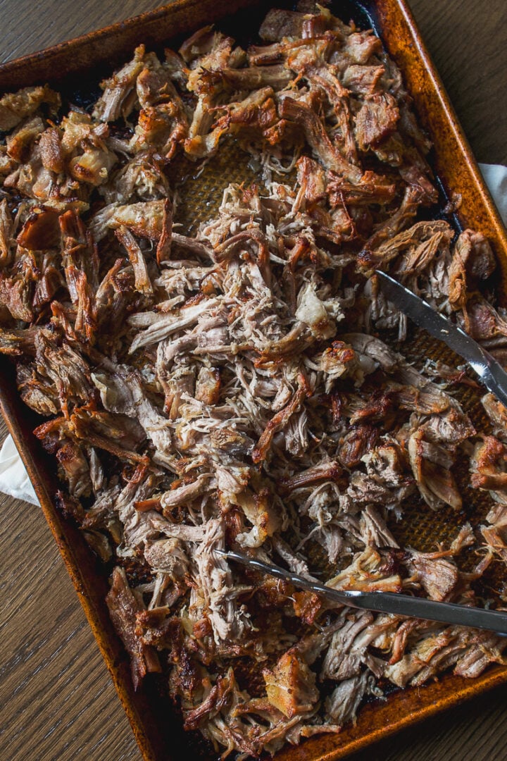 A metal baking sheet filled with shredded crockpot carnitas, with metal tongs resting on top.