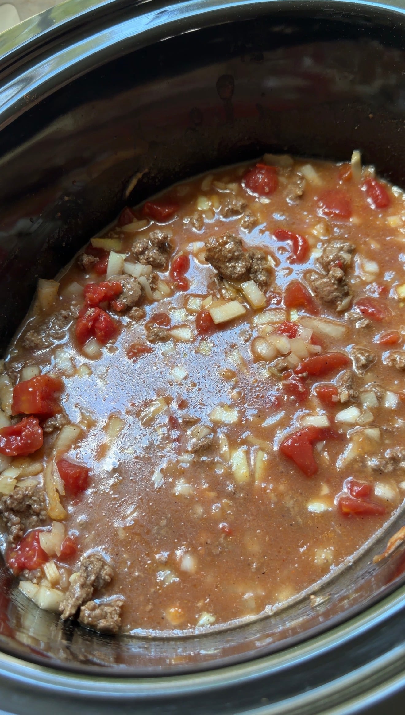 A slow cooker filled with ground meat, diced onions, and tomato chunks in a thin brown broth.