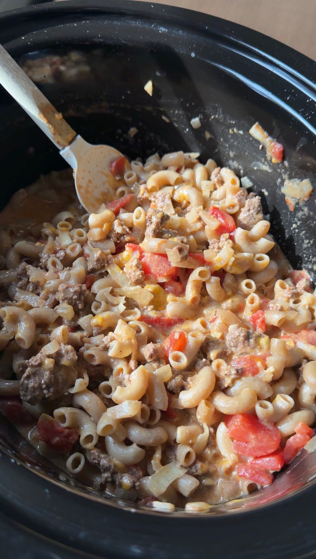 A slow cooker filled with creamy pasta, ground meat, and diced tomatoes, with a large serving spoon resting inside.