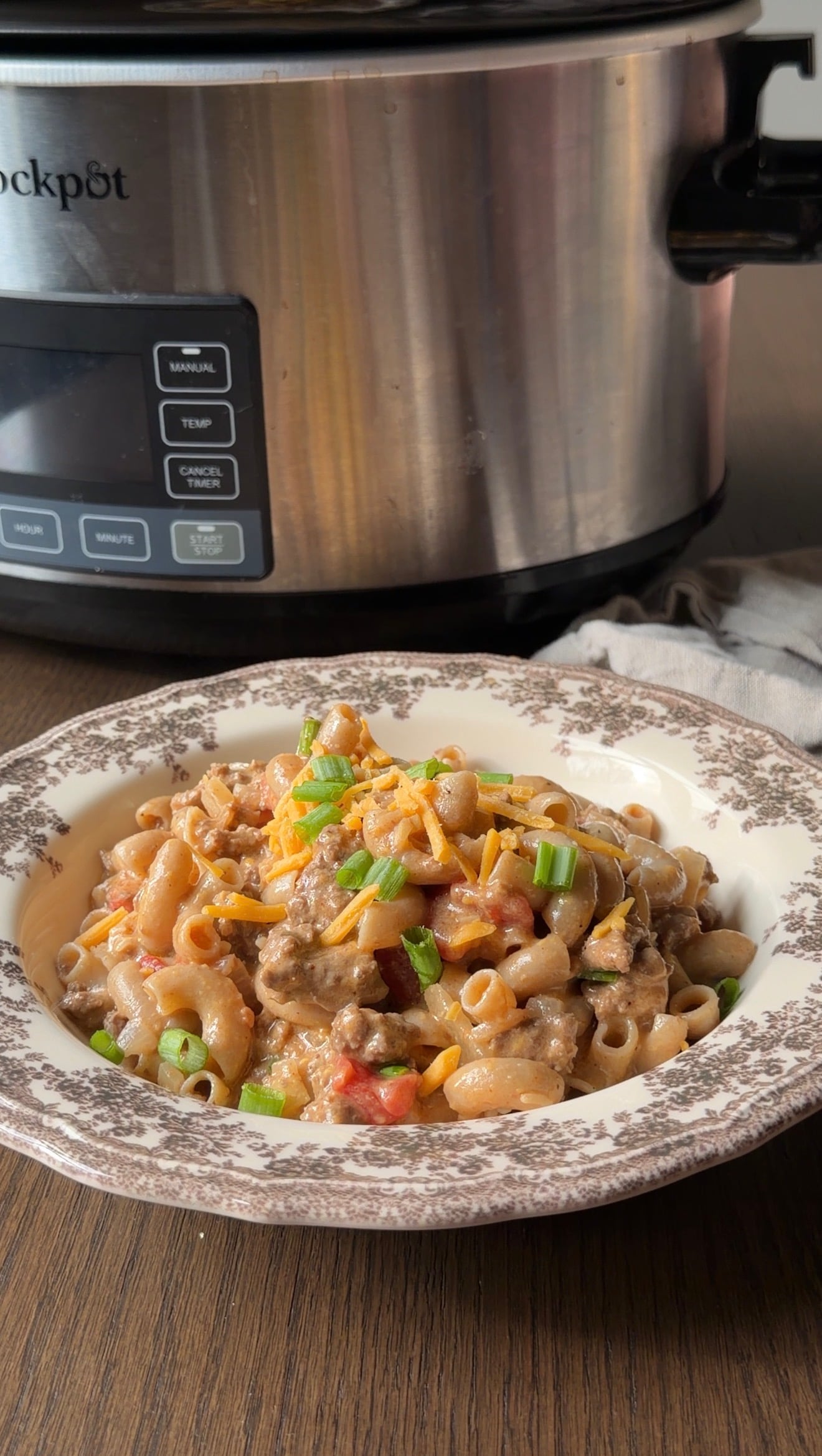 A bowl of cheeseburger macaroni topped with shredded cheddar cheese and green onions, sitting in front of a slow cooker.