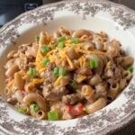 A bowl of creamy pasta with ground meat, topped with shredded cheddar cheese and chopped green onions.