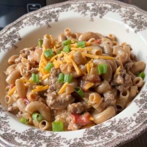A bowl of creamy pasta with ground meat, topped with shredded cheddar cheese and chopped green onions.
