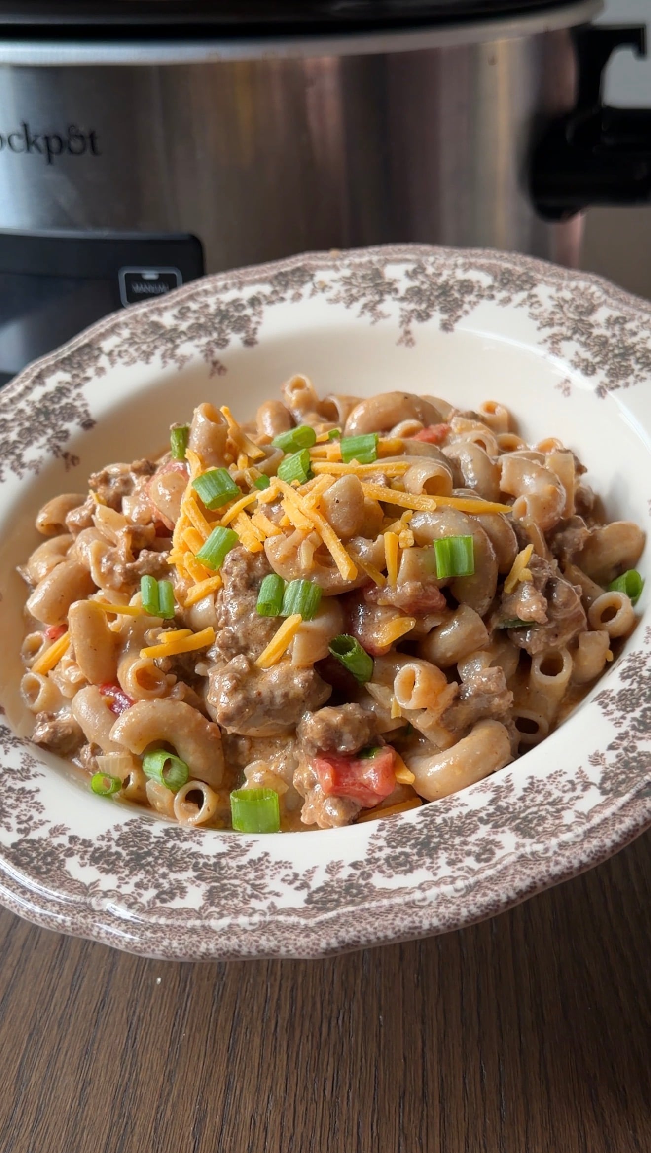 A bowl of creamy pasta with ground meat, topped with shredded cheddar cheese and chopped green onions.