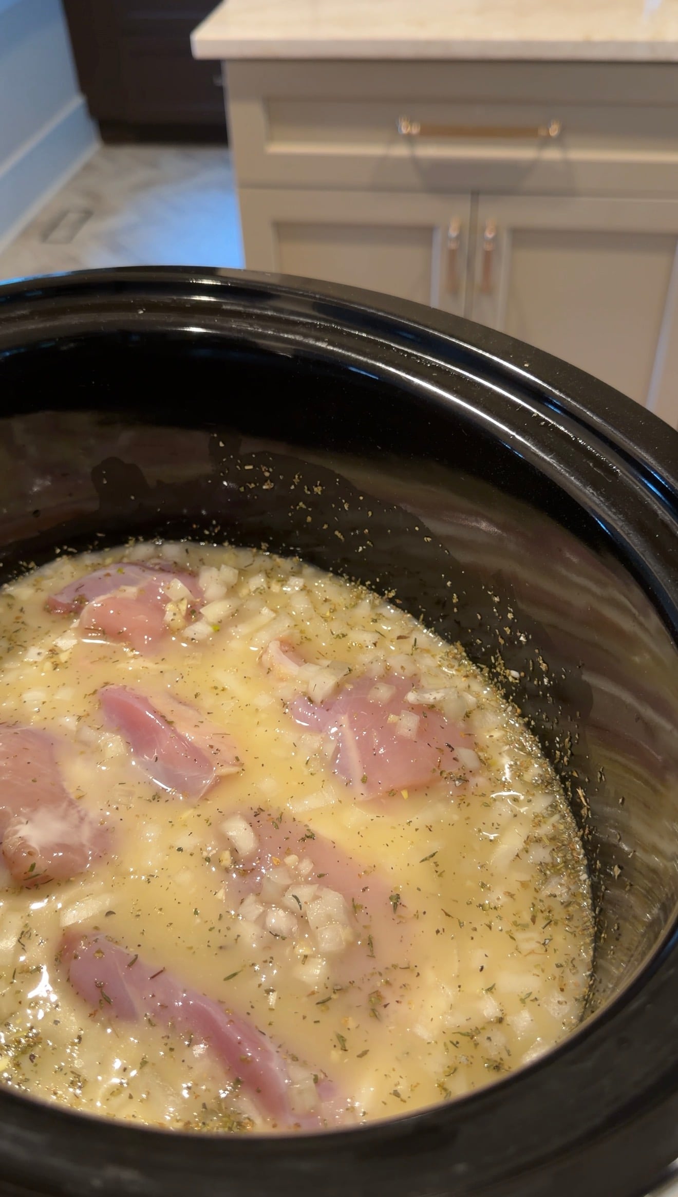 Raw chicken pieces, chopped onions, and herbs in a yellowish liquid are inside a black slow cooker, ready to be cooked. Kitchen cabinets and part of the floor are visible in the background.