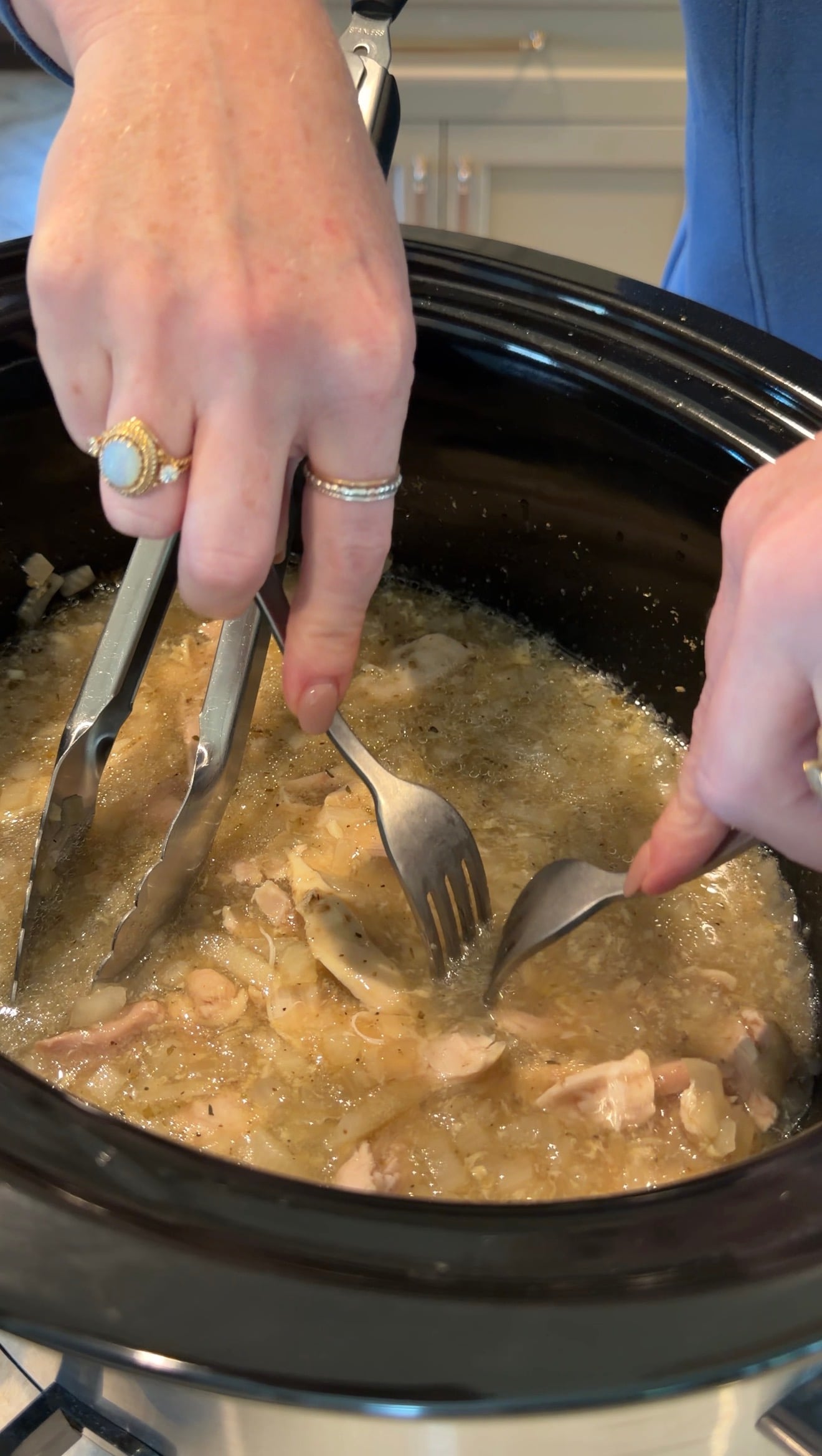Two hands use metal tongs and two forks to shred cooked chicken in a slow cooker filled with broth. The person wears rings on multiple fingers. The scene is focused on the food preparation process.