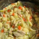 Close-up of a creamy chicken and gnocchi stew with carrots, peas, and celery, cooked in a black pot. A serving spoon is partly visible on the right side of the image.