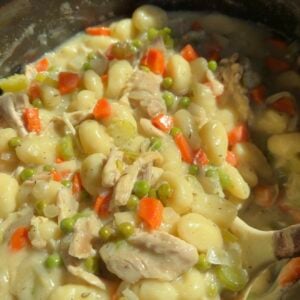 Close-up of a creamy chicken and gnocchi stew with carrots, peas, and celery, cooked in a black pot. A serving spoon is partly visible on the right side of the image.