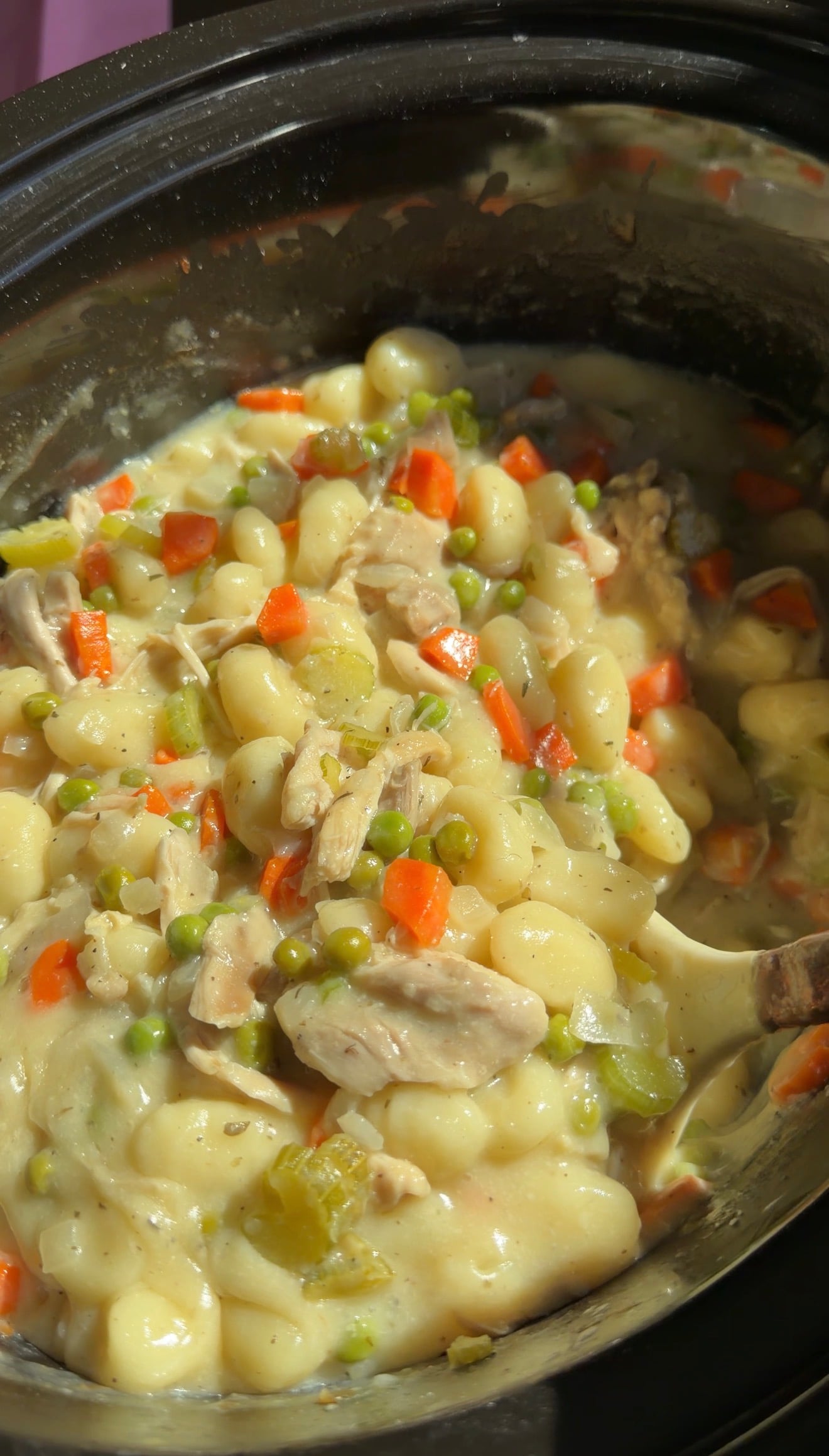 Close-up of a creamy chicken and gnocchi stew with carrots, peas, and celery, cooked in a black pot. A serving spoon is partly visible on the right side of the image.