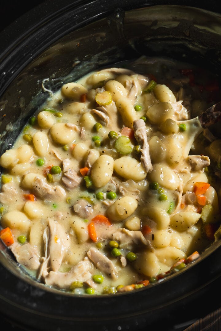 Creamy Crockpot Chicken Pot Pie Gnocchi stew with peas, carrots, and celery in a black slow cooker. This hearty dish is thick and full of tender vegetables and pillowy gnocchi.