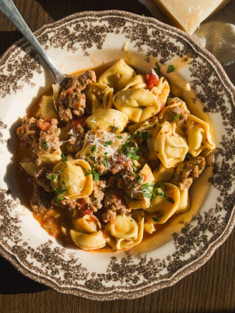 A bowl of tortellini with ground meat and tomato sauce, topped with grated cheese and herbs, with a spoon and a block of parmesan nearby.