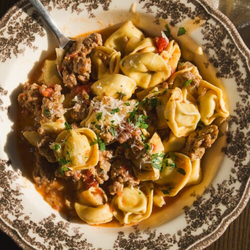 A bowl of tortellini with ground meat and tomato sauce, topped with grated cheese and herbs, with a spoon and a block of parmesan nearby.