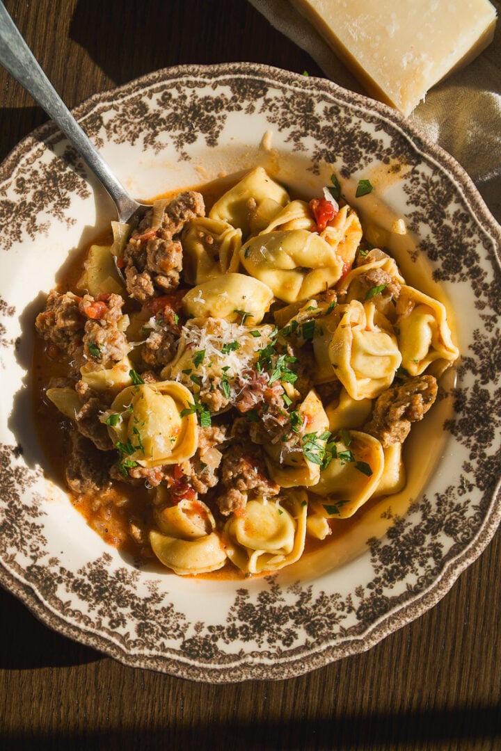 A bowl of tortellini with ground meat and tomato sauce, topped with grated cheese and herbs, with a spoon and a block of parmesan nearby.