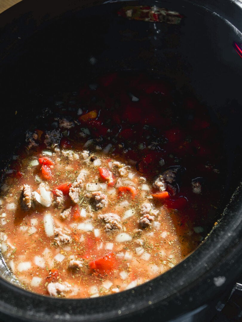 A slow cooker filled with a stew base of ground meat, chopped tomatoes, onions, and herbs in broth.