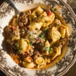 A bowl of tortellini with sausage and tomato sauce, topped with grated cheese and herbs, next to a wedge of parmesan.