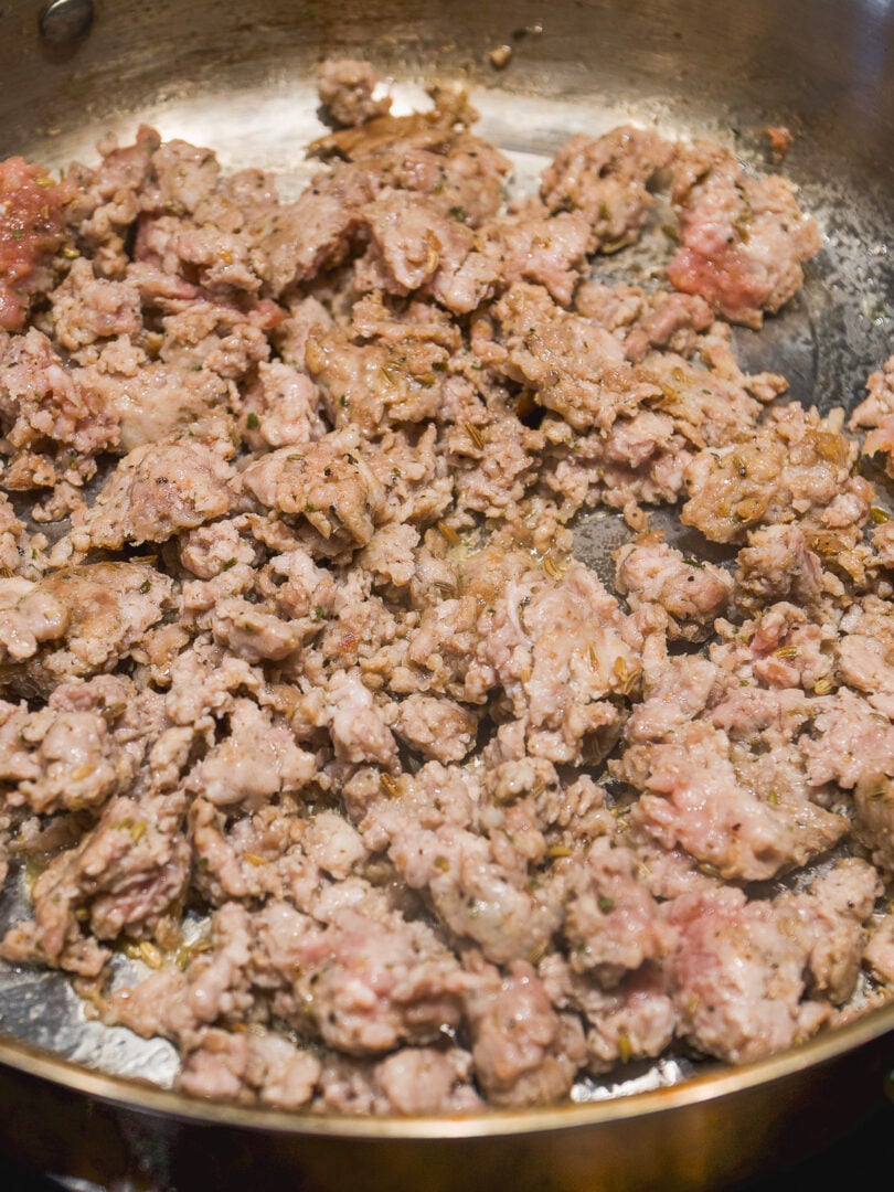 Ground meat with herbs browning in a stainless steel skillet.