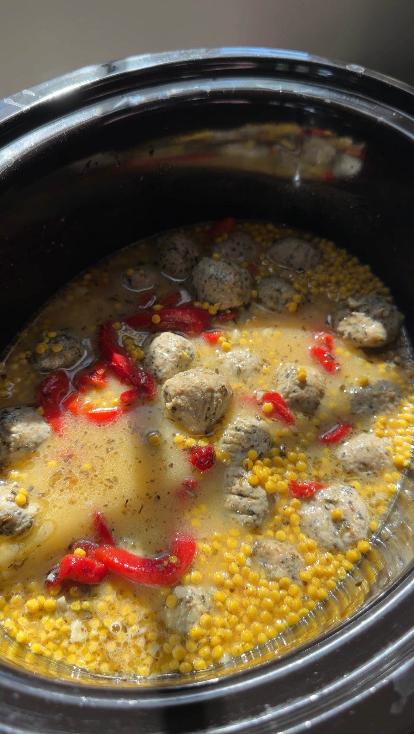 A slow cooker filled with meatballs, red bell pepper strips, and small yellow grains in a light broth.