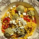 A bowl of pearl couscous mixed with red bell peppers, topped with Greek meatballs and crumbled feta cheese, with a silver spoon resting in the bowl.